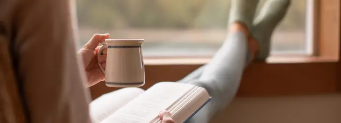 Person relaxing by a window with a mug in one hand and an open book in the other, wearing cozy socks.