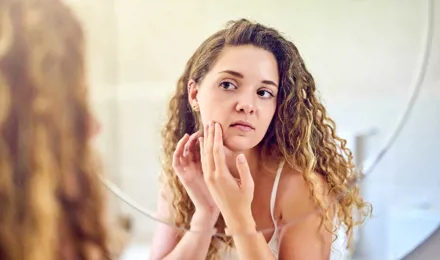 A young woman squeezing a pimple on her oily skin and looking at the mirror