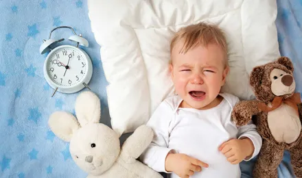 A baby crying on a blanket surrounded by two cuddly toys and an analogue alarm clock