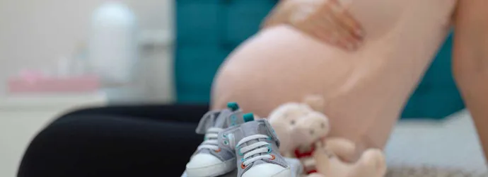 A neat pile of baby's clothes on a bed with a pregnant women sat in the background