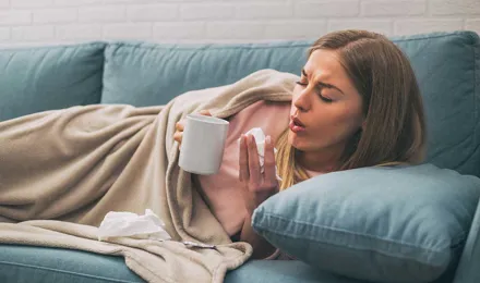 Woman lying in the sofa coughing while holding tea cup and a tissue