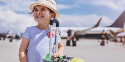 Young girl smiling with a plane in the background holding a yellow suitcase of items you need for a family holiday