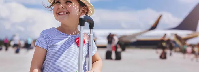 Young girl smiling with a plane in the background holding a yellow suitcase of items you need for a family holiday