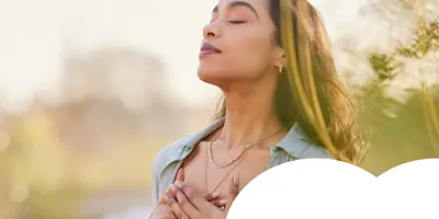 A woman stands outdoors with eyes closed and hands on her chest, taking a peaceful, mindful breath in a soft natural setting.