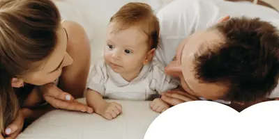 A mother and father lie on a bed facing their baby, smiling and interacting closely during a tender family moment.