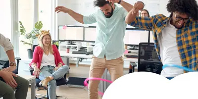 Group of adults in an office having fun, with two people hula-hooping and others laughing and cheering.
