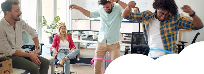 Group of adults in an office having fun, with two people hula-hooping and others laughing and cheering.
